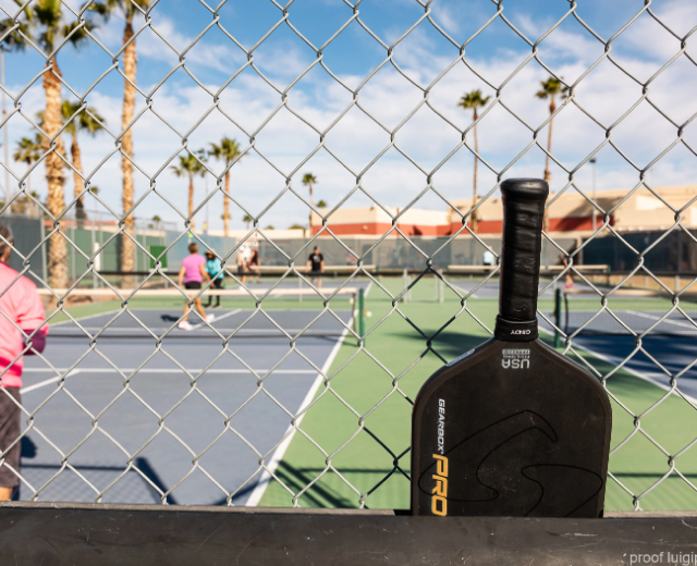 A pickleball racket leaning against a fence with several pickleball courts in the background.